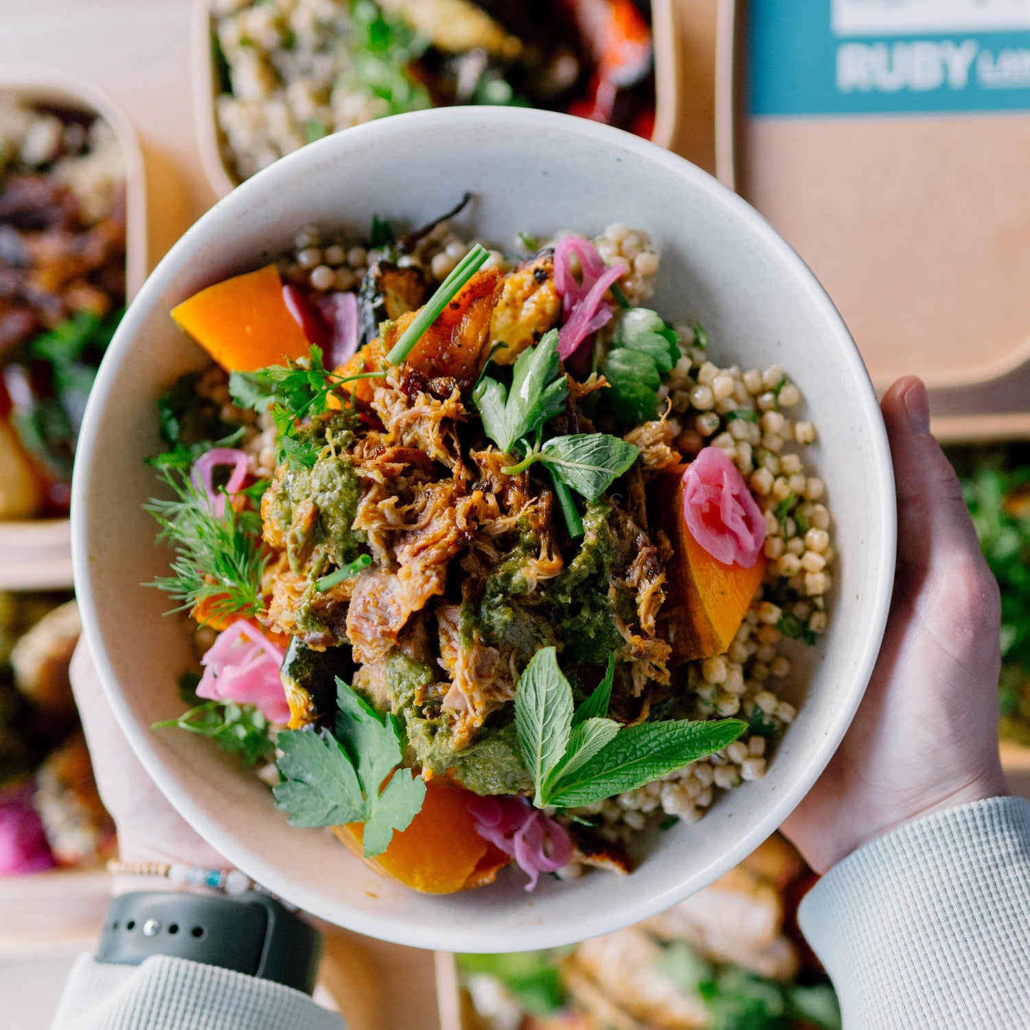 Person holding a bowl of salad with various ingredients, including vegetables and grains, with more bowls of food in the background.
